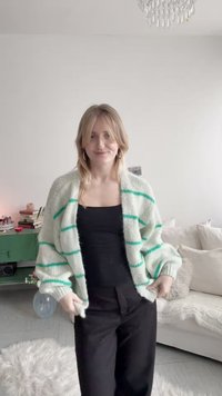 Mujer con cabello rubio que lleva un cárdigan blanco con rayas verdes sobre una blusa negra y pantalones negros, de pie en una sala iluminada y acogedora.