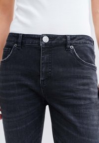 Schwarze Denim-Jeans mit glatter Textur, ausgestattet mit einem silbernen Knopfverschluss, fünf Taschen und einem dezenten Verblassen für einen getragenen Look.