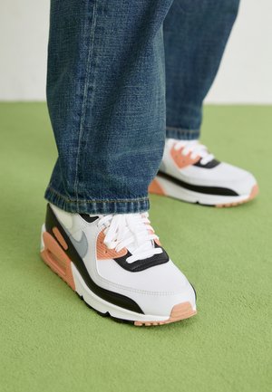 Persona que lleva zapatillas blancas, negras y durazno con cordones, de pie sobre alfombra verde, vestida con jeans de mezclilla azul.