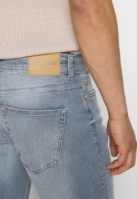 Jeans in denim azzurro chiaro con una finitura texturizzata. Presentano un'etichetta in pelle marrone chiaro sulla vita e una tasca posteriore con un contorno cucito.