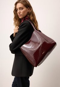 Bordo odinė rankinė su blizgiu krokodilo raštu, minkšta forma ir viena petneša, su auksiniu logotipo akcentu.