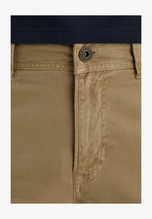 Beżowe bawełniane spodnie z zamknięciem na guzik z przodu, charakteryzujące się gładką teksturą i prostym designem. Widoczne szwy i zarysy kieszeni.