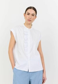 Camicia di cotone bianca con maniche corte arrotolate e un pannello arricciato sul davanti. Abbinata a pantaloni azzurri chiari. Texture liscia e design minimal.