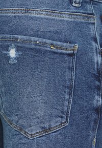 Back pocket of blue denim jeans with yellow stitching and slight distressed tear near upper left corner.