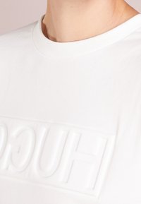 Camiseta blanca de algodón con un logo "HUGO" en relieve y texturizado en el pecho. Diseño clásico de cuello redondo con un acabado suave.