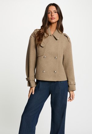 Frau trägt eine beige, zweireihige Wolljacke mit geknöpften Manschetten und dunkelblaue Jeans mit weitem Bein vor einem schlichten weißen Hintergrund.