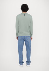 Felpa verde a maniche lunghe con polsini a coste, caratterizzata da un design a strisce nere sulla schiena, abbinata a jeans azzurri chiari e scarpe bianche.