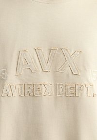 Beżowy sweter z podniesionym haftem tekstu "AVX" i "AVIREX DEPT." w subtelnym odcieniu kremowym. Krótkie rękawy, miękka struktura materiału.