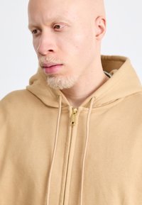 Tan hoodie in soft fabric, featuring a front zipper and drawstring hood. Smooth texture with golden zipper hardware.