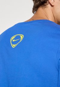 Plava pamučna majica s okruglim izrezom i kratkim rukavima, s žutim Nike logotipom na gornjem dijelu leđa. Glatka tekstura, ležeran dizajn.