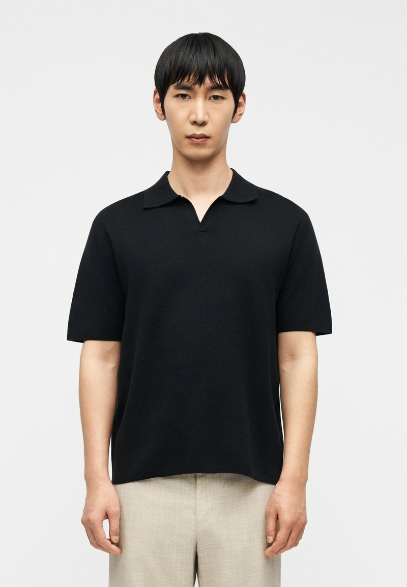 Νέος άνδρας με ίσια μαύρα μαλλιά, που φοράει απλό μαύρο κοντομάνικο πουκάμισο με γιακά και ανοιχτόχρωμο μπεζ παντελόνι, μπροστά σε λευκό φόντο.