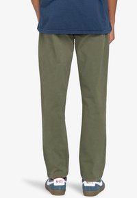 Pantaloni in cotone verde con una vestibilità rilassata, gambe dritte e tasche laterali, abbinati a sneakers blu e bianche con logo.