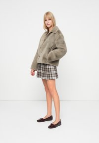 Veste oversized en fausse fourrure de couleur vert olive atténué, présentant un grand col et une fermeture à boutons, assortie à une mini-jupe à carreaux et des chaussures sombres brillantes.