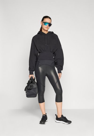 Fekete cropped kapucnis pulóver zsinórral, fényes fekete leggings és fekete sportcipők. Kiegészítők közé tartozik egy fekete edzőtáska és kék napszemüveg.