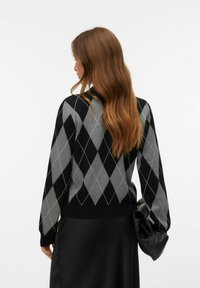 Svart tröja med grått rutmönster, tydlig V-hals och ribbade muddar. Kombinerad med en svart kjol och håller en svart clutchväska.