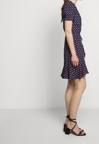 Donna in piedi con un abito a motivi geometrici, maniche a sbuffo e sandali neri con cinturino alla caviglia e tacco largo, su uno sfondo uniforme.