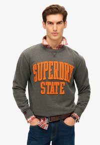 Szary sweter z dużym pomarańczowym napisem "SUPERDRY STATE". Długie rękawy, okrągły dekolt, nałożony na czerwoną i białą koszulę w kratę. Niebieskie dżinsy.