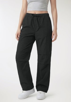 Donna che indossa pantaloni cargo neri larghi con tasche laterali, un top corto grigio chiaro e scarpe da ginnastica bianche, in piedi davanti a uno sfondo bianco.