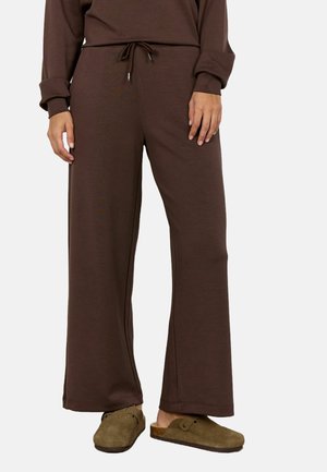 Persona con pantalones marrones holgados con cordón y zapatos tipo mocasín verde oliva, de pie delante de un fondo claro y sencillo.