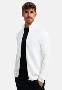 Weiße Zip-Jacke mit strukturiertem Fischgrätmuster, geripptem Kragen und Bündchen, kombiniert mit einem schwarzen Shirt darunter.