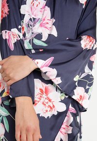 Granatowa sukienka w kwiaty z różowymi i białymi motywami. Posiada długie dzwonowe rękawy i gładką teksturę materiału. Widoczny detal poniżej łokcia.