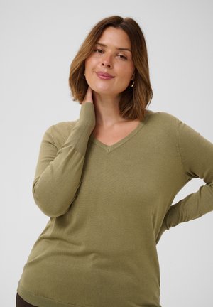 Femme aux cheveux bruns mi-longs portant un pull marron clair à col en V, touchant son cou et regardant doucement la caméra.