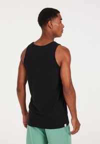 Canotta nera in cotone con spalline larghe, vestibilità regolare e schiena bassa. Indossata con pantaloni shorts verdi chiari. Dettaglio logo discreto sull'orlo.