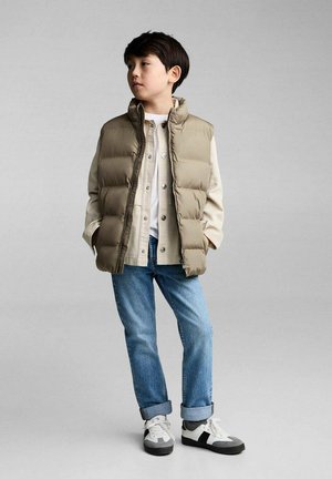 Beige polstret vest over en lys skjorte, parret med blå jeans. Vesten har høj krave og er fyldig i teksturen med sidelommer.