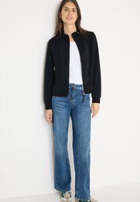 Černá bunda na zip s vysokým límcem, doplněná modrými širokými džínami a šedými teniskami. Bunda má nenápadné zipy a přiléhavý střih.