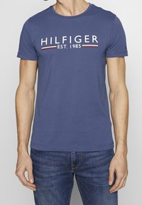 T-shirt di cotone blu navy con colletto rotondo, maniche corte e scritta bianca "HILFIGER EST. 1985" con strisce rosse e bianche. Vestibilità standard.