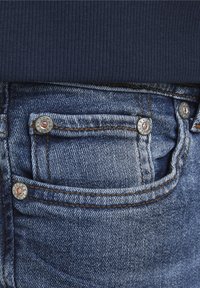 Dunkelblaues, geripptes Gewebe überlappt den Bund von verblassten blauen Jeans. Bemerkenswerte Details sind Messingschrauben und kontrastierende orangefarbene Nähte.