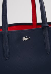 Sac cabas bleu marine texturé avec doublure intérieure rouge et logo crocodile argenté sur le devant, avec deux bretelles.