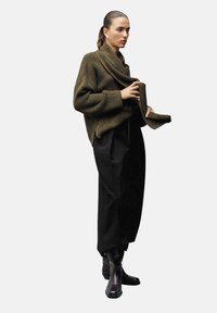 Suéter de punto de gran tamaño en verde oliva con un escote drapeado, combinado con pantalones negros de tiro alto y botas negras de altura hasta el tobillo.