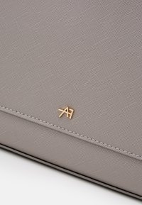 Szary materiał o teksturze z delikatnym wzorem, z małym złotym logo. Smukły design z przeszyciem i wypolerowanymi krawędziami.