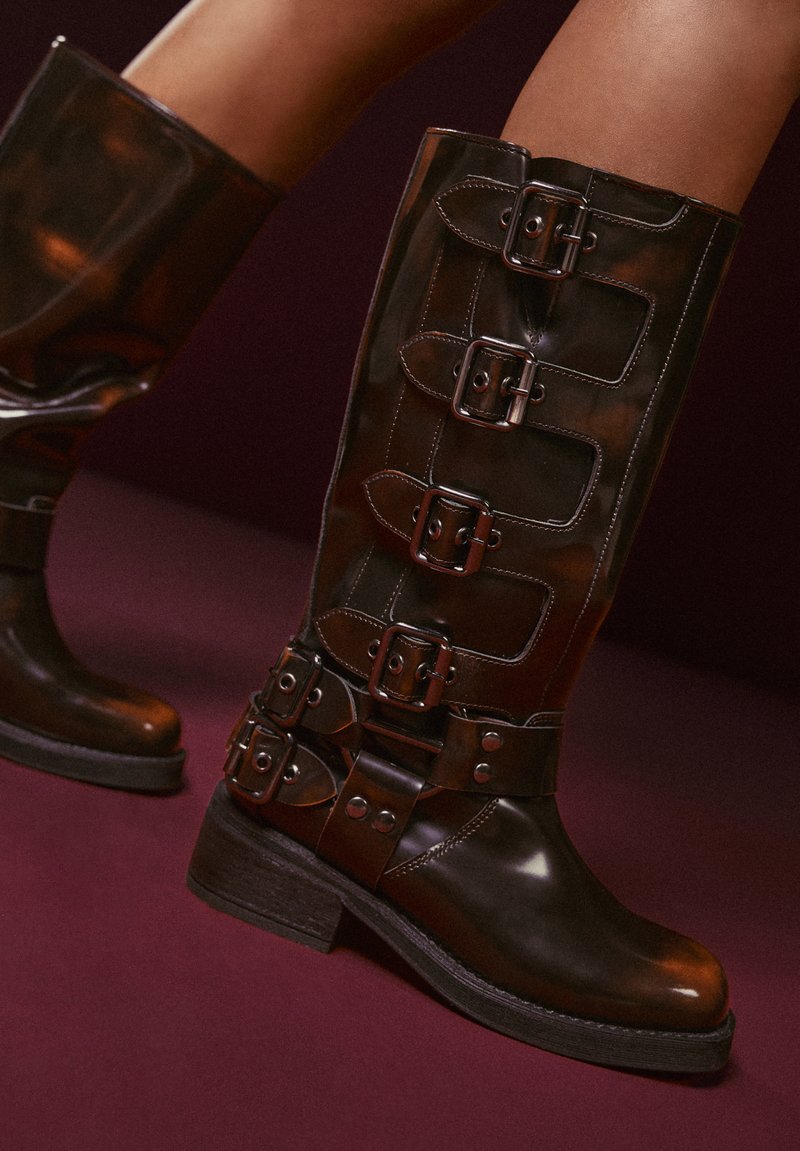 Stivali alti fino al ginocchio in pelle marrone con più fibbie d'argento, punta arrotondata e tacco a blocco robusto. Texture lucida con dettagli cuciti.