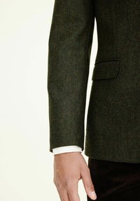 Grün gemusterte Tweedjacke im Fischgrätmuster mit taillierter Passform. Verfügt über eine Seitentasche, einen Knopfdetail und einen sichtbaren weißen Shirtmanschette darunter.