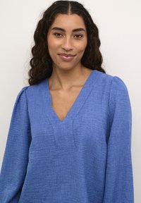 Blouse à manches longues, col en V en tissu bleu texturé. Présente des manches légèrement bouffantes et une coupe décontractée avec des coutures sur le devant.