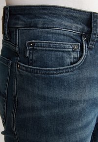 Dunkelblaue Jeans aus Denim mit schmaler Passform, ausgestattet mit einer kleinen Münztasche, Metallnieten und dezenter Ausbleichung entlang des Beins.