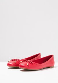 Ballerines en cuir rouge avec un bout arrondi, présentant un accent de nœud à l'avant et une semelle en caoutchouc plate. Texture lisse et design minimal.