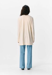 Lungo cardigan crema con texture a coste, caratterizzato da orlo dritto e spalle scese, abbinato a jeans a gamba larga blu chiaro e scarpe nere.