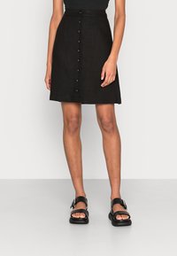 Schwarzer ausgestellter Rock aus strukturiertem Stoff mit Knopfleiste. Trägt dazu schwarze Sandalen mit dicker Sohle und Schnallendetail.