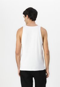 Débardeur en coton blanc avec un col rond et des bretelles larges, porté avec un pantalon noir, vu de l'arrière. Texture lisse.