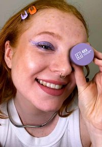 Lila kosmetikbehållare märkt "423" hålls bredvid ett leende ansikte med silver smycken. Har pastellmakeup och texturerat, vågigt hår.
