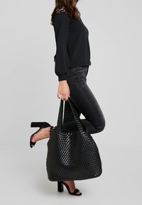 Bolso tote negro de tejido con un acabado texturizado, que cuenta con dos asas resistentes y un diseño amplio, combinado con ropa negra y tacones.
