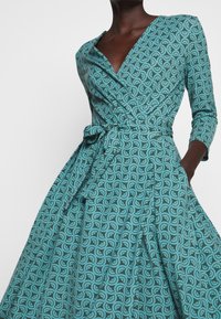 Robe portefeuille bleu sarcelle à motif circulaire, manches trois-quarts, encolure en V, taille ajustée avec lien et jupe plissée avec poches.