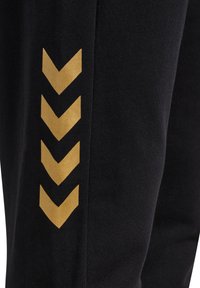 Schwarze Jogginghosen mit glatter Textur, ausgestattet mit einem goldenen Zickzackmuster auf der linken Seite. Einfaches Design, elastischer Bund und schmal zulaufende Passform.