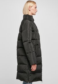 Cappotto imbottito nero con colletto alto, cuciture orizzontali, cerniera frontale e tasche laterali. Materiale morbido e lucido con silhouette lunga.