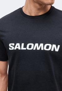 Μαύρο μπλουζάκι με λευκό λογότυπο SALOMON, φορεμένο από άτομο που φαίνεται από τον λαιμό μέχρι το μέσο του κορμού.