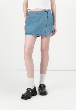 Persoon die een blauwe denim wikkel mini-rok, witte sokken, zwarte schoenen met gesp en een wit cropped top draagt tegen een witte achtergrond.