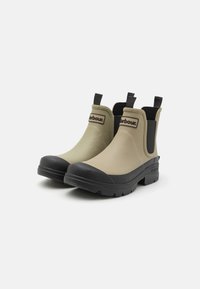 Beige rubber ankle boots with black soles, elastic side panels, and pull tabs. Features a black logo on the front. Textured sole for grip.
